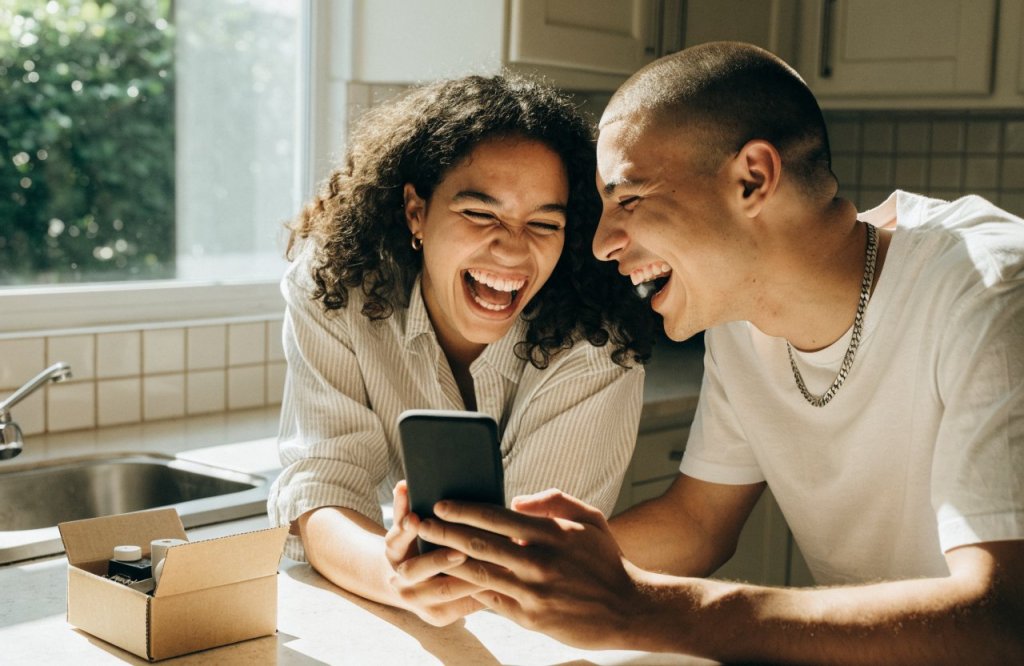 Dois amigos no balcão da cozinha rindo enquanto olham o celular, com uma caixa aberta ao lado