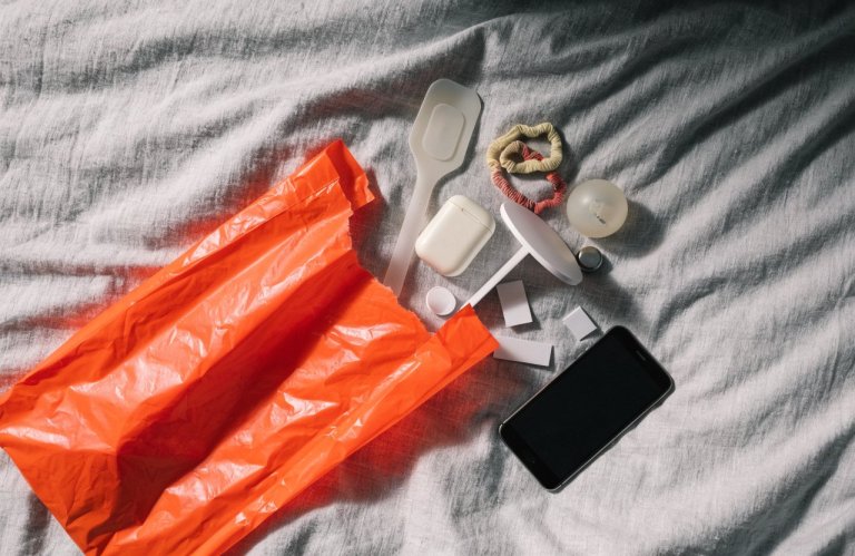 Embalagem laranja aberta sobre a cama com pequenos produtos e um celular ao lado